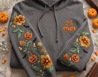 May include: A charcoal gray hoodie featuring intricate embroidery on both sleeves and the chest. The sleeves display orange jack-o'-lanterns, yellow flowers, green leaves, small black bats, and candy corn. The chest features two orange jack-o'-lanterns tied with a bow.