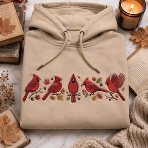 Op de afbeelding: Een beige hoodie met een borduursel van vijf rode kardinaalvogels op takken, omringd door kleine kleurrijke herfstbladeren en bessen, voorzien van bijpassende beige trekkoorden bij de halslijn.