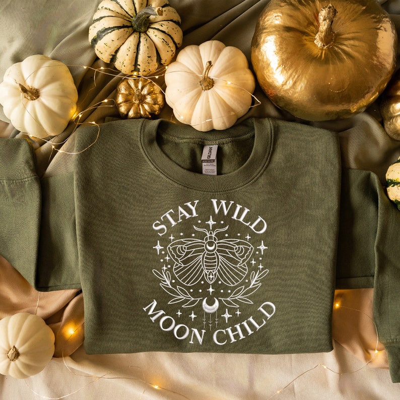 Puede incluir: Una sudadera verde con un dise&ntilde;o gr&aacute;fico blanco que presenta una polilla, estrellas y el texto "Stay Wild Moon Child".