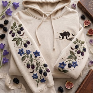 Könnte beinhalten: Ein cremefarbener Kapuzenpullover mit aufwendigen Stickereien an beiden Ärmeln und auf der Brust. Die Ärmel zeigen Brombeerranken, blaue Glockenblumen, kleine Fledermäuse und Sternbilder. Auf der Brust ist eine schwarze Katze auf einem Besen gestickt.