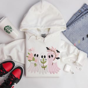 May include: A white hooded sweatshirt featuring an embroidered design of three cartoon ghosts holding flowers and branches. The ghosts are in shades of pink and white. The image also includes a pair of black and red sneakers, a Starbucks cup, and blue jeans.