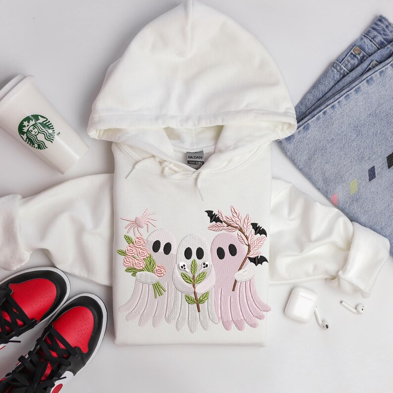May include: A white hooded sweatshirt featuring an embroidered design of three cartoon ghosts holding flowers and branches. The ghosts are in shades of pink and white. The image also includes a pair of black and red sneakers, a Starbucks cup, and blue jeans.