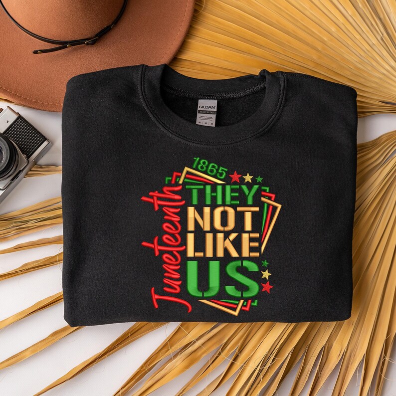 May include: Black sweatshirt with a green, red, and gold graphic design that says "Juneteenth 1865 They Not Like Us" with stars.