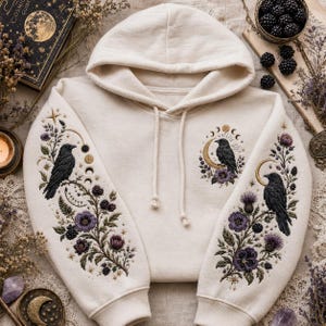 May include: An off-white hooded sweatshirt featuring detailed embroidery on both sleeves and the chest. The design includes black ravens perched on branches, purple flowers, blackberries, and golden moon phases, creating a cohesive botanical theme.