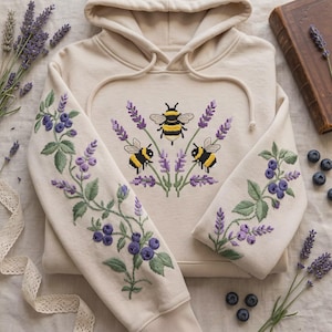May include: A beige hooded sweatshirt featuring detailed embroidery of three bumblebees surrounded by purple lavender sprigs on the chest. The sleeves are decorated with embroidered green vines, purple lavender, and clusters of blue blueberries.