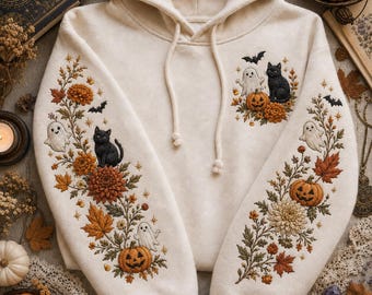 May include: A cream-colored hooded sweatshirt featuring intricate embroidery on the sleeves and chest. The design includes black cats, white ghosts, orange pumpkins, autumn leaves, and small bats in a detailed seasonal arrangement.