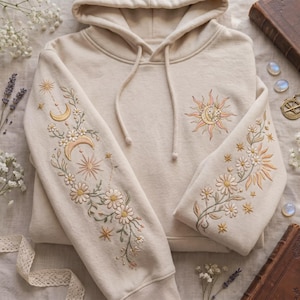 Op de afbeelding: Een beige hoodie met gedetailleerd borduurwerk op beide mouwen en op de borst. Het ontwerp bevat hemelse motieven zoals zonnen, maansikkels en sterren, verweven met delicate bloemranken en bloemen die op madeliefjes lijken.