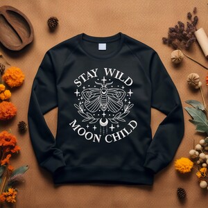 Puede incluir: Sudadera negra con un dise&ntilde;o gr&aacute;fico blanco que presenta una polilla, estrellas y el texto "Stay Wild Moon Child".