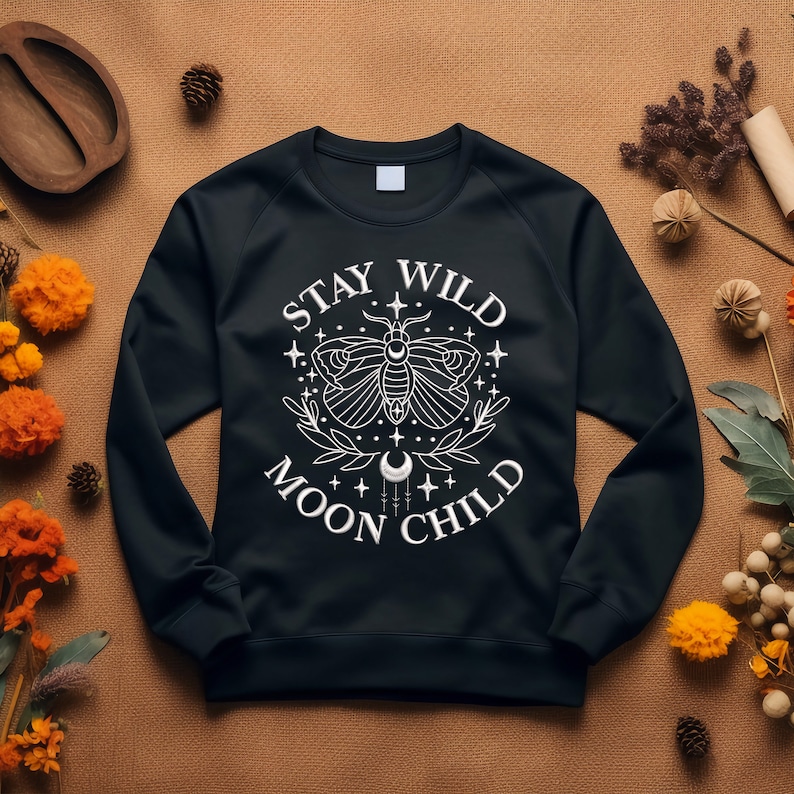 Puede incluir: Sudadera negra con un dise&ntilde;o gr&aacute;fico blanco que presenta una polilla, estrellas y el texto "Stay Wild Moon Child".