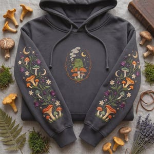 Könnte beinhalten: Ein anthrazitfarbener Kapuzenpullover mit aufwendigen Stickereien. In der Mitte ist ein grüner Frosch auf einem Pilz zu sehen, während beide Ärmel mit einem botanischen Muster aus bunten Pilzen, Blumen und Mondsicheln verziert sind.