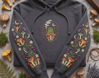 May include: A charcoal gray hooded sweatshirt featuring intricate embroidery. The center displays a green frog sitting on a mushroom, while both sleeves are decorated with a botanical pattern of colorful mushrooms, flowers, and crescent moons.