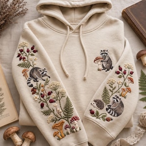May include: A beige hooded sweatshirt featuring detailed embroidery on both sleeves and the chest. The designs include raccoons, a hedgehog, various mushrooms, ferns, and berry sprigs. The hoodie has a drawstring hood and long sleeves.