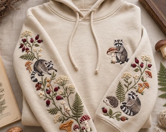 May include: A beige hooded sweatshirt featuring detailed embroidery on both sleeves and the chest. The designs include raccoons, a hedgehog, various mushrooms, ferns, and berry sprigs. The hoodie has a drawstring hood and long sleeves.