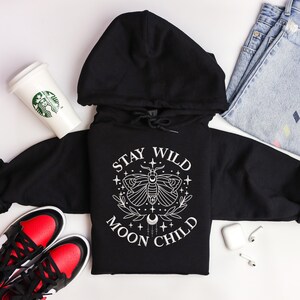 Puede incluir: Sudadera con capucha negra con un dise&ntilde;o gr&aacute;fico blanco que presenta una polilla, una luna creciente y el texto "Stay Wild Moon Child".