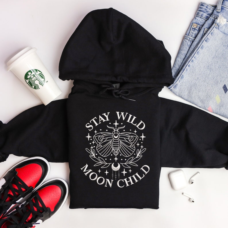 Puede incluir: Sudadera con capucha negra con un dise&ntilde;o gr&aacute;fico blanco que presenta una polilla, una luna creciente y el texto "Stay Wild Moon Child".