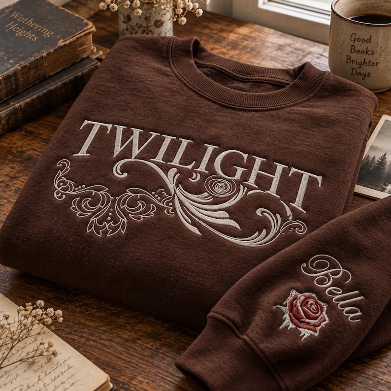 May include: A dark brown crewneck sweatshirt featuring the word TWILIGHT embroidered in white across the chest with decorative white scrollwork. The sleeve features the name Bella embroidered in white above a red rose with green leaves.