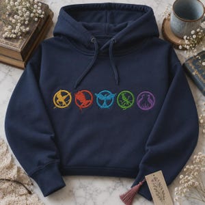 May include: A navy blue hooded sweatshirt featuring a row of five circular embroidered designs in yellow, red, light blue, green, and purple. Each circle contains a distinct bird-like silhouette. The hoodie has long sleeves and a drawstring hood.