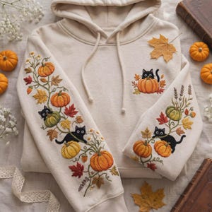 Peut inclure: Un sweat à capuche couleur crème orné de broderies détaillées représentant des chats noirs, des citrouilles orange et des feuilles d'automne. Le motif s'étend sur les manches et inclut un petit chat sortant d'une citrouille sur la poitrine.