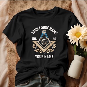 May include: Black t-shirt featuring a Masonic emblem with the text "YOUR LODGE NAME" above and "YOUR NAME" below. The emblem includes a compass, square, and the letter "G". The number "56" is also displayed.