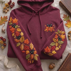 Peut inclure: Un sweat-shirt à capuche bordeaux orné de broderies détaillées de feuilles d'automne et de petits chats noirs. Les manches présentent une vigne de feuillage orange, jaune et vert avec des têtes de chats, et un motif similaire orne la poitrine.