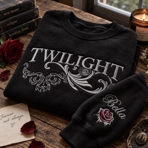 May include: A black crewneck sweatshirt featuring white embroidered text that reads TWILIGHT with ornate swirling floral patterns underneath. The sleeve includes an embroidered red rose and the name Bella in white cursive script lettering.