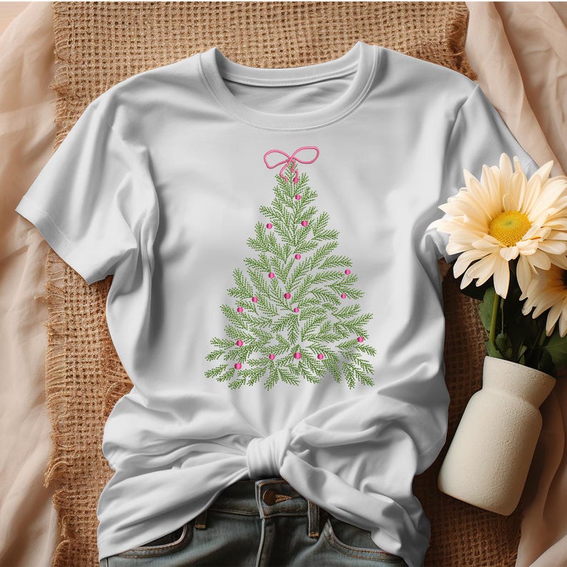 May include: White t-shirt featuring an embroidered Christmas tree design. The tree is green with pink ornaments and a pink bow at the top. The shirt is made of a soft material and is perfect for the holiday season.