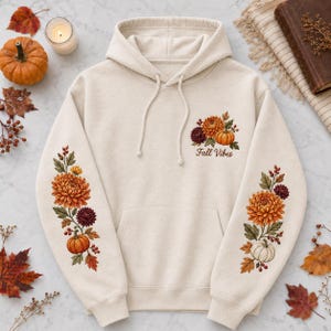 May include: A cream-coloured hooded sweatshirt featuring a front pocket and drawstrings. The chest and sleeves are decorated with floral illustrations of orange and dark red chrysanthemums, berries, and pumpkins, with the text Fall Vibes.