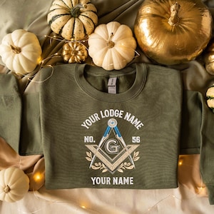 May include: An olive green sweatshirt with the text "YOUR LODGE NAME" and "YOUR NAME" in white. The design includes a compass, square, and the letter "G". The number "56" is also visible. The sweatshirt is surrounded by pumpkins.