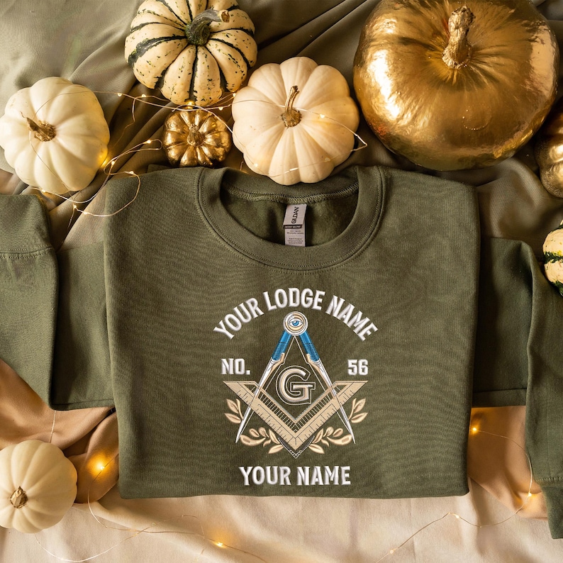 May include: An olive green sweatshirt with the text "YOUR LODGE NAME" and "YOUR NAME" in white. The design includes a compass, square, and the letter "G". The number "56" is also visible. The sweatshirt is surrounded by pumpkins.