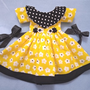 May include: A yellow doll dress with white floral print and a black polka dot collar. The dress has a black hem and is adorned with two black polka dot bows. The dress is approximately 10 inches in length.