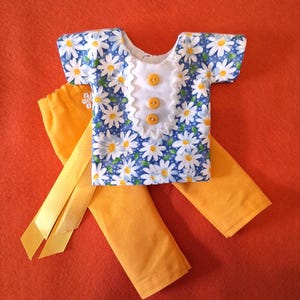 May include: A short-sleeved shirt featuring a blue background with a white daisy pattern, a white center panel with ruffled edges, and three yellow buttons. Paired with solid yellow pants, the outfit includes a yellow ribbon with white dots.