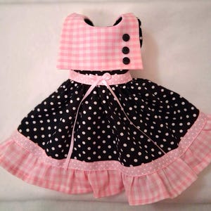 May include: A doll-sized dress with a pink and white gingham top, black polka dot skirt, and pink ribbon accents. The dress features a ruffled hem and three black buttons. The overall design is reminiscent of a vintage style.