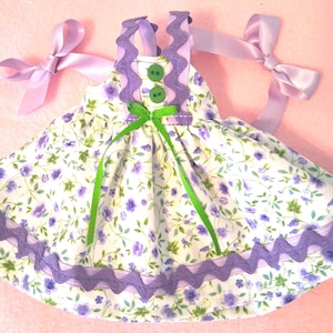 May include: A miniature dress with a floral pattern of purple flowers and green leaves on a white background. The dress has purple trim, green ribbon accents, and lavender bows on the straps. Two green buttons adorn the bodice.