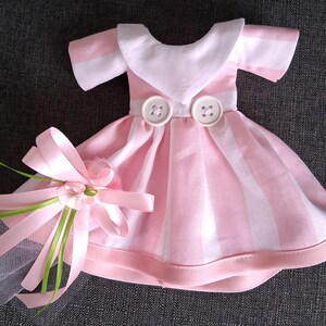May include: A doll-sized pink and white striped dress with a white collar and two large buttons. A pink bow with green accents and a small flower is attached to the dress, suggesting a special occasion or decorative item.