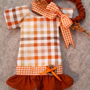 May include: A short-sleeved dress featuring a white, orange, and brown plaid pattern. The dress has a solid rust-colored ruffled hem, a matching orange gingham bow, and a small wooden button accent. A coordinating rust-colored elastic headband with an orange gingham bow and small orange berry sprigs is attached.