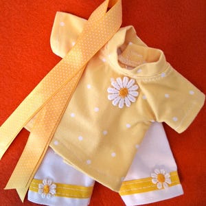 May include: A yellow short-sleeved shirt with white polka dots and a white daisy applique on the chest, paired with white pants featuring yellow ribbon trim and small daisy appliques, accompanied by a long yellow ribbon with white polka dots.