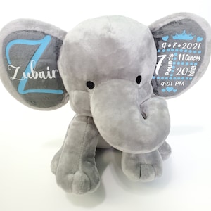 May include: Gray plush elephant stuffed animal with embroidered details. One ear has the name "Zubair" in blue lettering. The other ear has the date "4.7.2021", "7 Pounds", "11 Ounces", "20 Inches", and "4:01 PM" in black lettering.