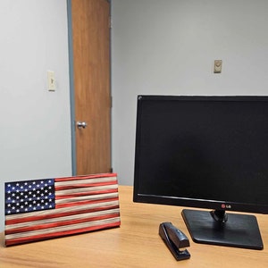 May include: A wooden American flag sits on a wooden desk next to a black computer monitor with the LG logo. A black stapler is also on the desk.
