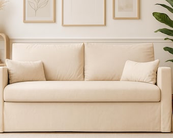 Op de afbeelding: Een crèmekleurige bank met twee grote rugkussens, twee kleinere rechthoekige sierkussens en een rokbasis. De bekleding is een effen, lichtbeige stof met een gladde textuur die de zitting, armleuningen en rugleuning volledig bedekt.