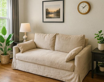 Op de afbeelding: Een beige loveseat met een losse hoes, voorzien van twee grote rugkussens, één zitkussen en twee kleine rechthoekige sierkussens. De bank heeft een rok en is bekleed met een gestructureerde stof in een neutrale kleur met strakke lijnen.