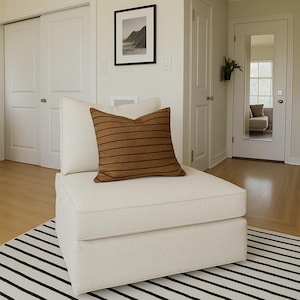 Puede incluir: Un sillón color crema con un cojín a rayas marrones. La silla está sobre una alfombra a rayas blancas y negras. Una fotografía de paisaje en blanco y negro enmarcada cuelga de la pared. La habitación tiene pisos de madera clara y puertas blancas.