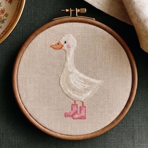 May include: Cross-stitch embroidery of a white goose wearing pink boots, set in a wooden hoop. The goose has an orange beak and pink cheeks. The embroidery is on a beige fabric, with a dark green background.