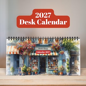 May include: A 2027 desk calendar featuring a watercolor-style illustration of a bookstore storefront with shelves of books, striped awnings, and potted plants. The top of the calendar displays the text 2027 Desk Calendar in white on a red background.