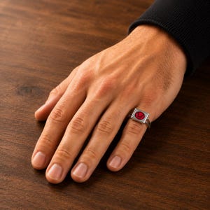 May include: A silver-toned ring featuring a square-shaped face with a red circular inlay containing a black graphic symbol. The ring is worn on the ring finger, showcasing a metallic band and a flat, geometric design with a bold colour contrast.