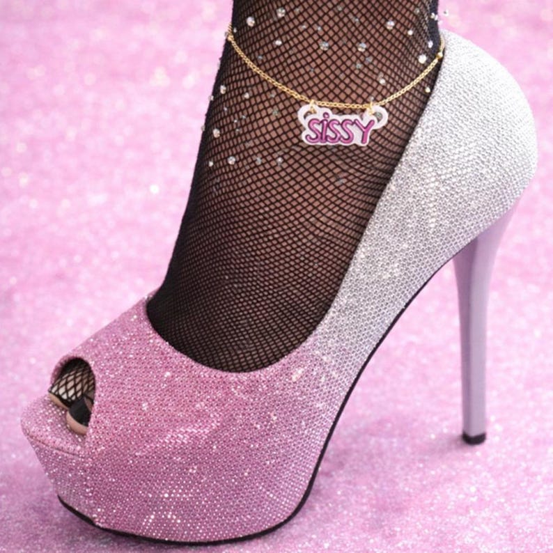May include: A gold-toned chain anklet featuring a white pendant with the word SISSY written in pink script. The anklet is worn with black fishnet stockings adorned with small rhinestones and a glittery pink and silver peep-toe high heel pump.