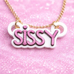 May include: A gold-tone chain necklace featuring a white acrylic pendant with the word SISSY in glittery pink lettering. The pendant has a small pink heart above the letter I and is attached to the chain with two gold-tone jump rings.