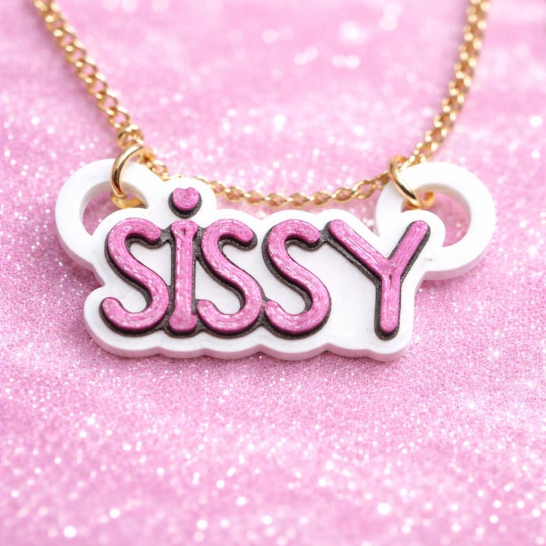 May include: A gold-tone chain necklace featuring a white acrylic pendant with the word SISSY in glittery pink lettering. The pendant has a small pink heart above the letter I and is attached to the chain with two gold-tone jump rings.