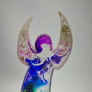 Può includere: Una statuetta di angelo in resina colorata con ali iridescenti e una base in legno. L'angelo è illuminato dal basso con una luce LED bianca.