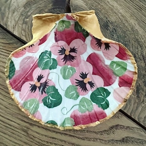 May include: A decorative seashell with a gold-painted edge. The interior is adorned with a floral pattern featuring pink and red pansies, green leaves, and white background. The shell is resting on a wooden surface.