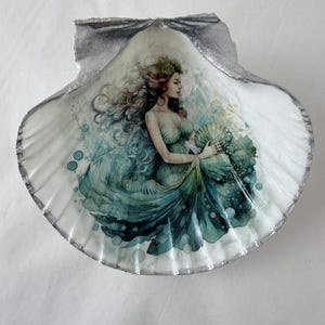May include: A decorative seashell featuring a watercolor painting of a mermaid. The mermaid is depicted in shades of green and blue, with long flowing hair and a flowing gown. The shell has a silver-painted edge.